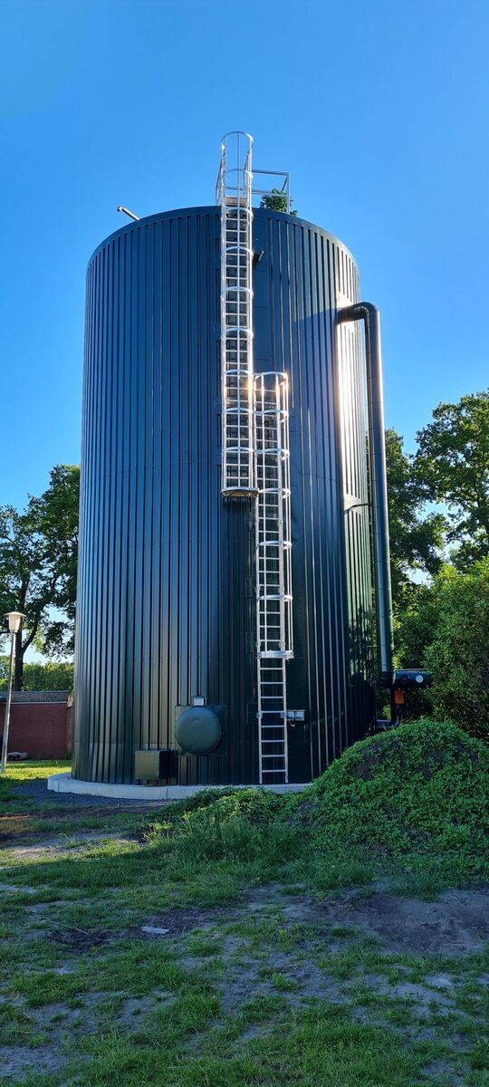 Ein großer, dunkelgrüner, zylindrischer Silo mit Leiter, umgeben von Bäumen bei klarem Himmel.