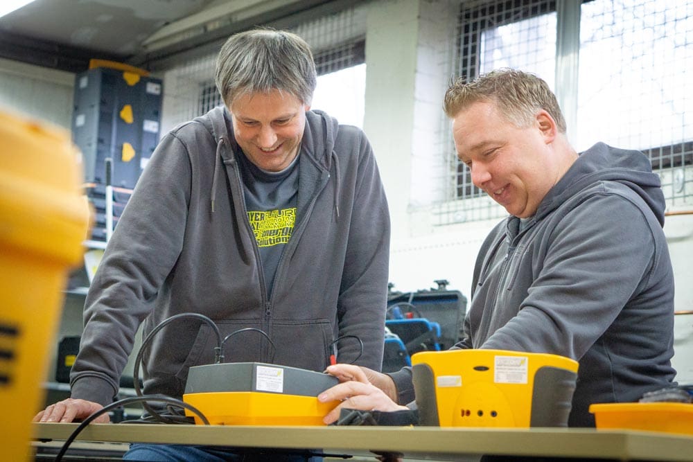 Zwei Männer mit grauen Hoodies lächeln über ein technisches Gerät auf einem Tisch.