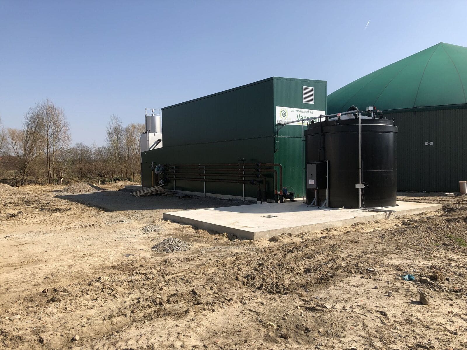 Biogasanlage mit grünem Gebäude, schwarzen Tanks und kahlen Bäumen im Hintergrund.