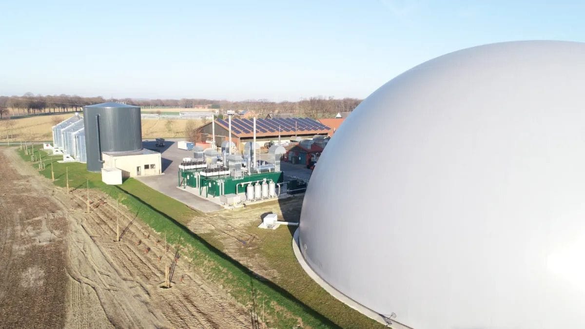 Biogasanlage mit grauem Kuppeltank, blauem Himmel und grüner Landschaft im Hintergrund.